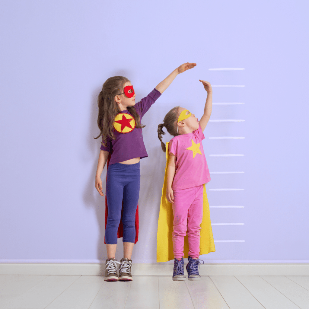 Two young girls stand side by side while comparing their height measurements against a wall chart. They are dressed as super heroes.