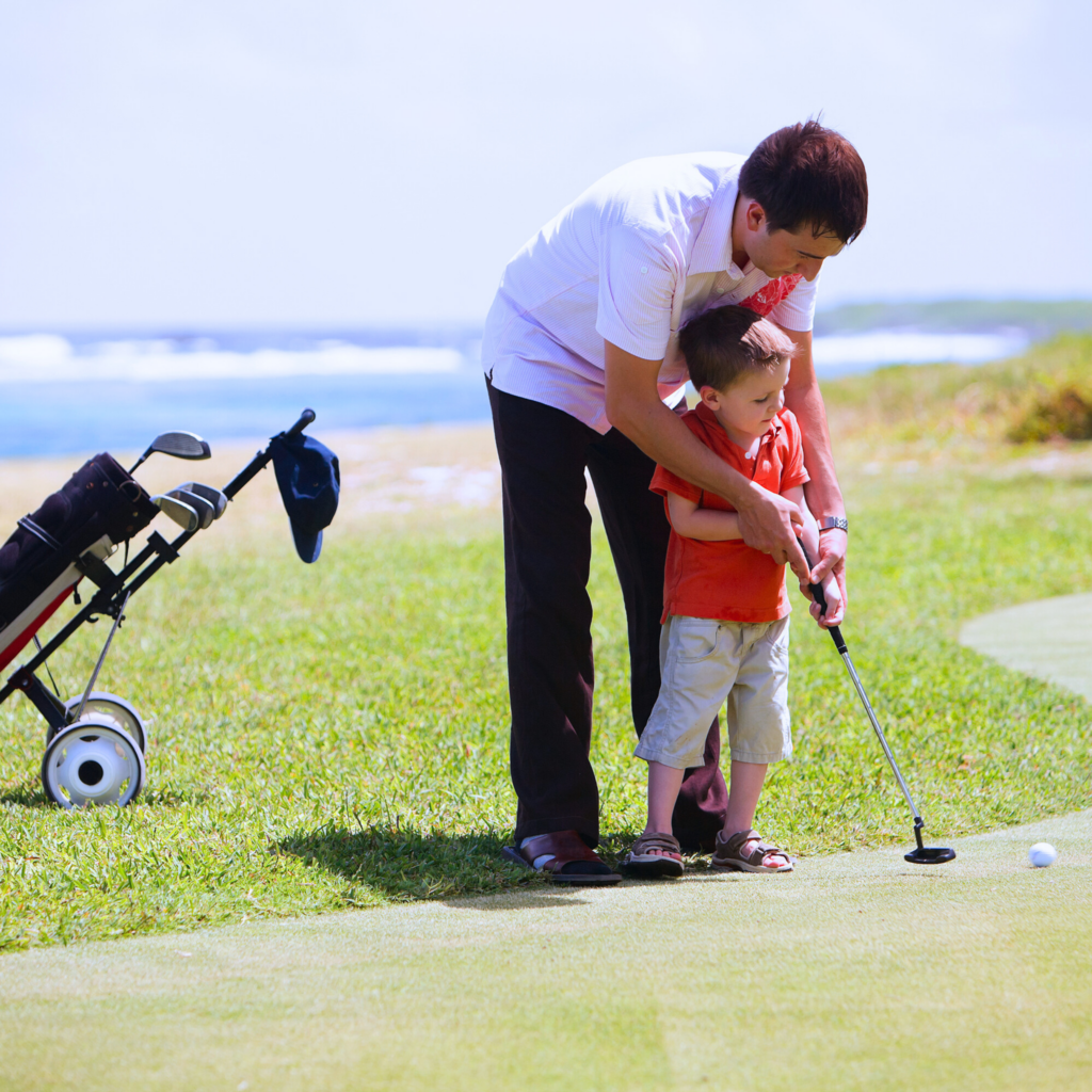 A man and a male child stand on a golf green in front of an ocean view. The man stands behind the child and is guiding him to hold a golf club. They are about to hit a golf ball together. To their right is a golf bag full of clubs.