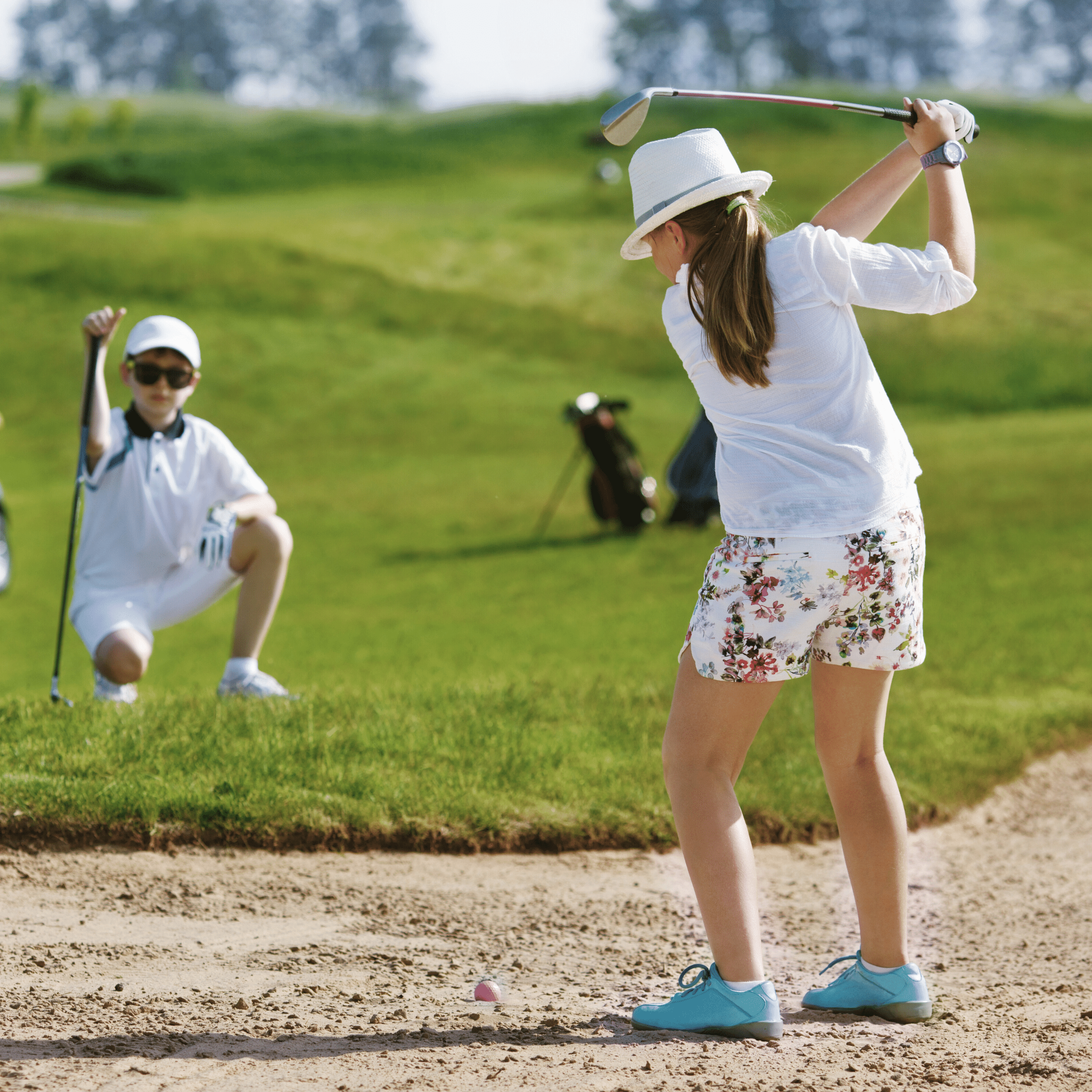 A female child stands in a golf sand bunker facing away. She is holding a golf club aloft and preparing to hit a golf ball. A male child is crouched on the grass in the background watching her shot.